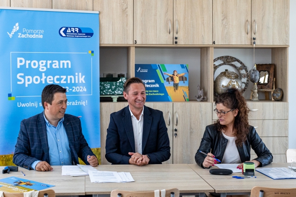 Podpisanie umowy dotacyjnej, w tle meble i niebieski roll-up